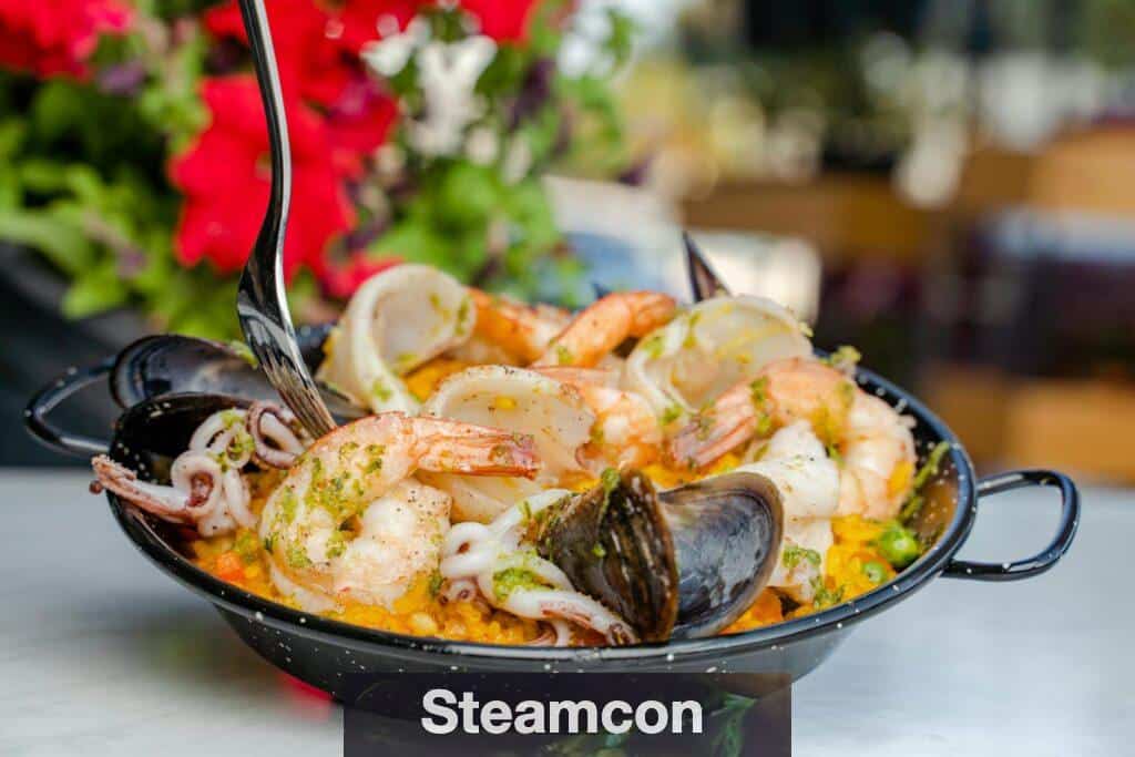 Close-up of a seafood paella dish in a black pan, garnished with shrimp, mussels, and calamari, served outdoors.